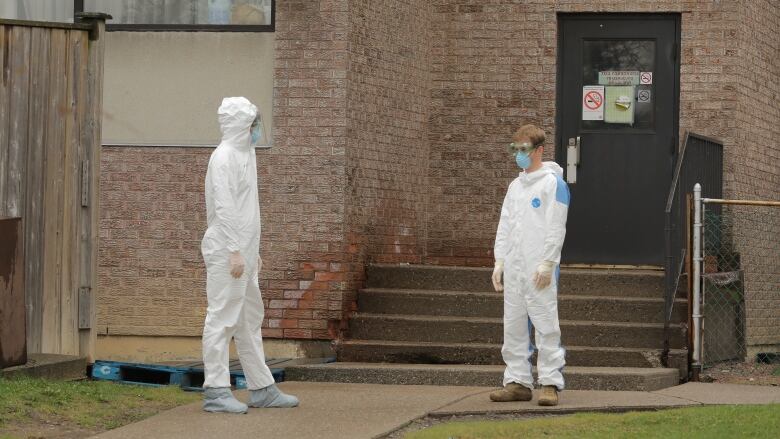 People in white hazmat suits stand outside of a brick building. People in white hazmat suits stand outside of a brick building.