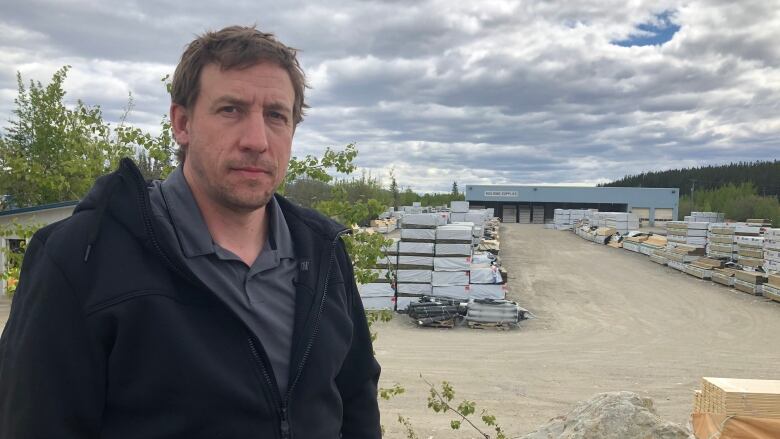 A man in a black rain jacket stands in front of a construction supply depot 