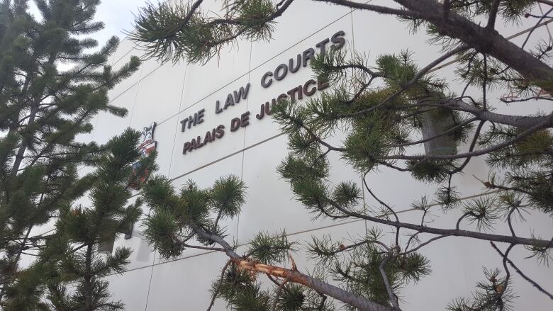 A sign on the side of a white tile building reads THE LAW COURTS PALAIS DE JUSTICE. There are tree branches in front. 