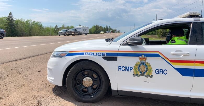 An officer in a fluorescent green vest sits in a white RCMP car at the side of a highway as traffic drives past.