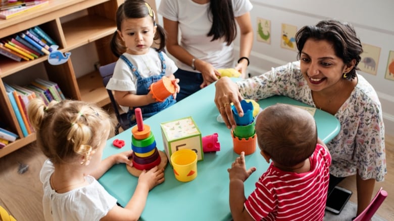 Three young children sit around a small table with two adults in a colourful daycare setting.