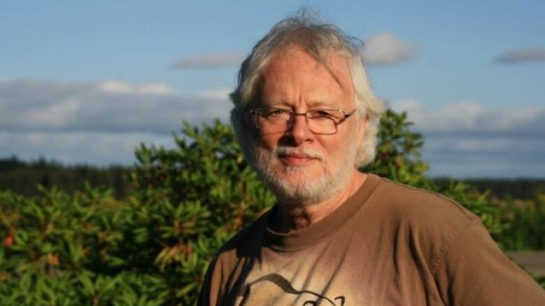 A man with grey hair and a grey beard wears T-shirt in front of foliage and blue skies. 