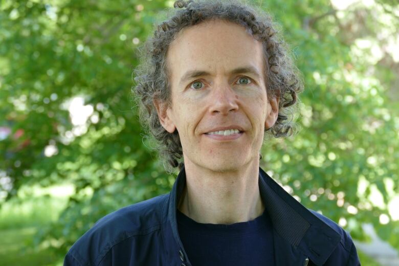 A portrait of a man with dark curly hair standing outside.