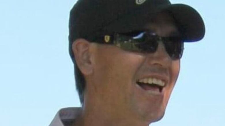 A man wearing sunglasses and a baseball hat with a broad smile. 