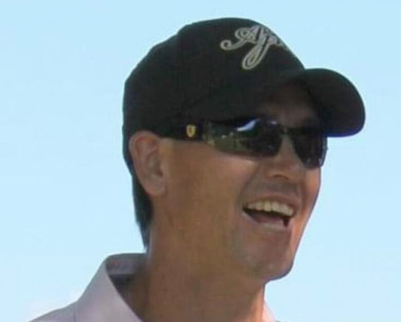 A man wearing sunglasses and a baseball hat with a broad smile. 