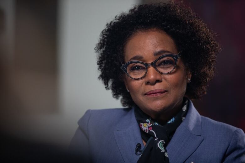 A Black woman with dark, curly hair wearing glasses, a blue blazer and a cravat