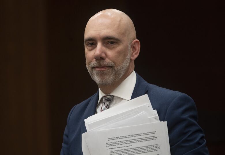 A bald man with a white grey beard who's wearing a blue suit holds a pile of papers.