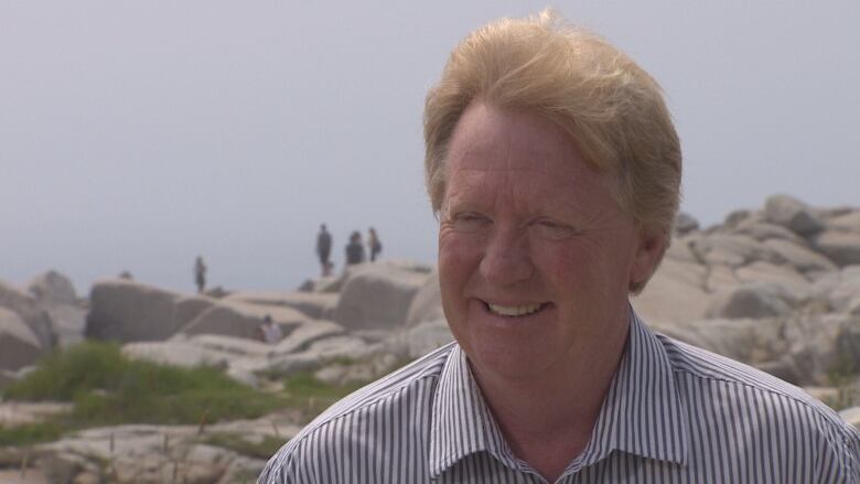 A white man with red hair and a striped shirt stands outside with people standing on white rocks behind him