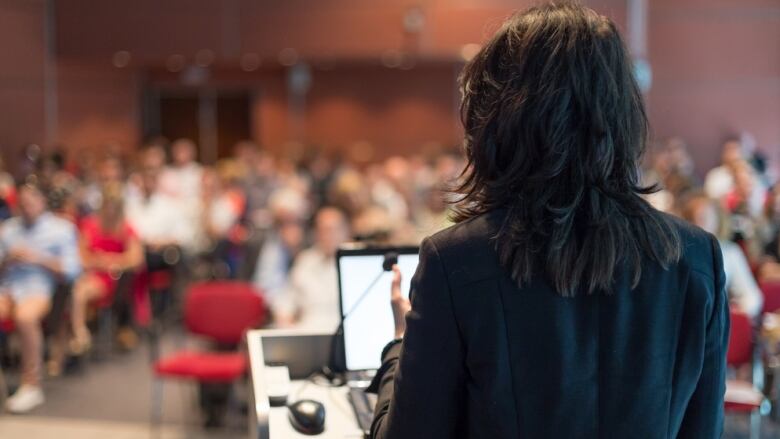 A woman with her back to the camera faces students in a lecture hall.