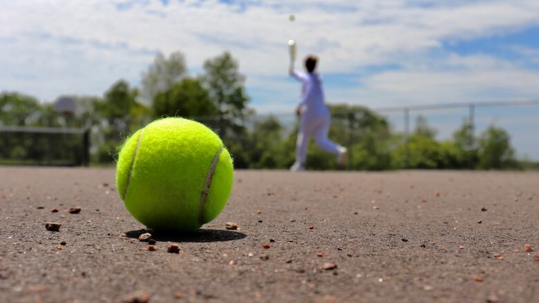 A tennis ball lays on the ground. There's a tennis player in the background.