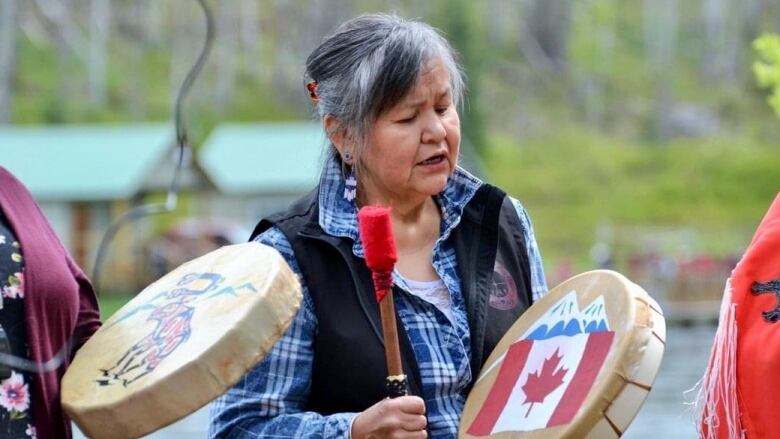 An Indigenous woman plays a drum and sings with standing with other people.