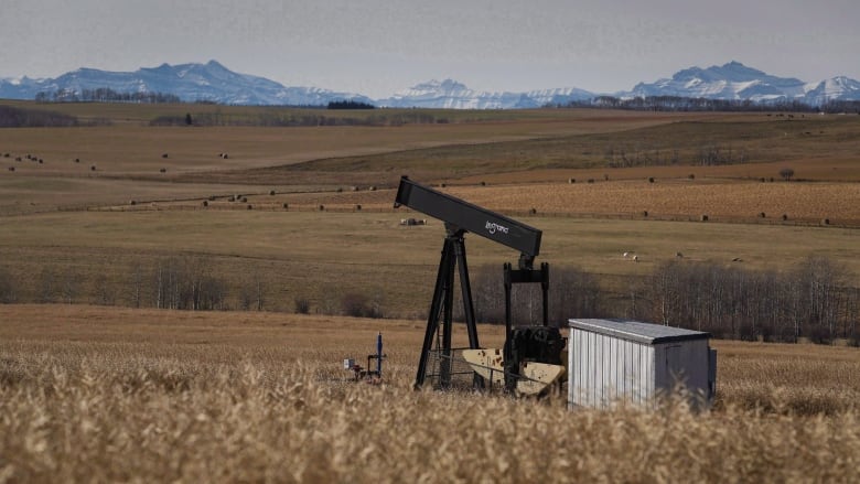 a prairie field is pictured at fall time, with an oil pumbjack in the middle and mountains in the horizon