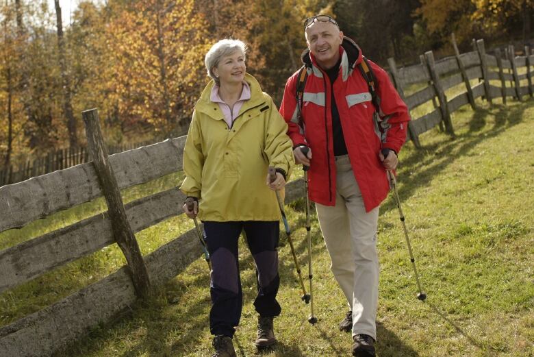 Two people walk with walking sticks along a wooden fence.