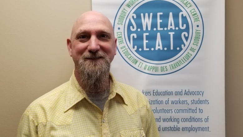 A bald man with a flowing beard in a yellow shirt stands in front of a sign saying the Sudbury Workers Education and Advocacy Centre