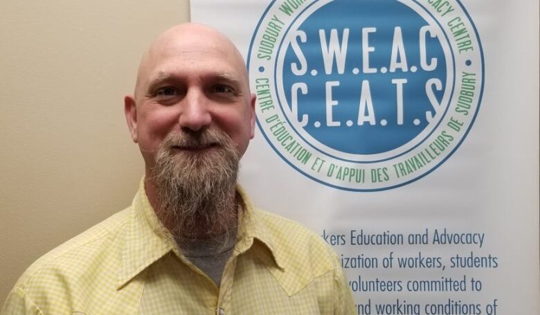 A bald man with a flowing beard in a yellow shirt stands in front of a sign saying the Sudbury Workers Education and Advocacy Centre