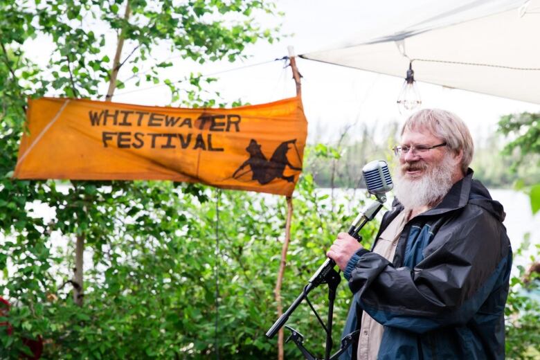 A man stands outdoors in front of a mic. He is smiling and talking to a crowd that is not pictured. 