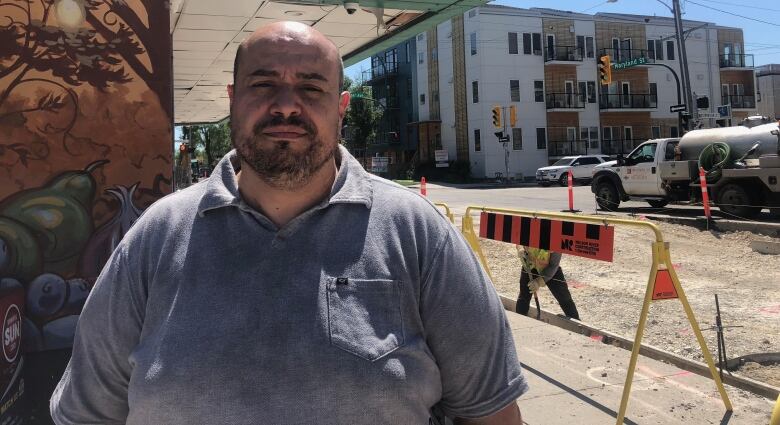 A man wearing a grey shirt stands on a sidewalk in front of  a building.