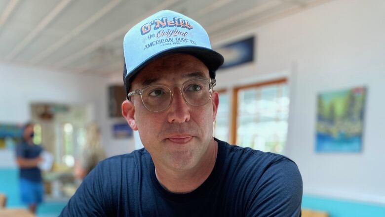 A man in a blue shirt, light blue hat and glasses sits at a table in a small restaurant.