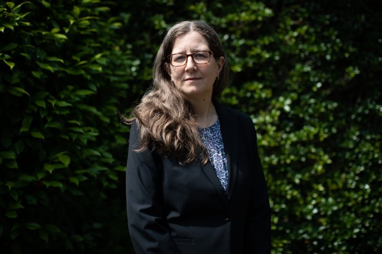 A woman stands outside, in front of a hedge.