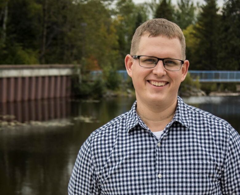 A man in a checkered shirt smiles, standing outside with a pond in the background.