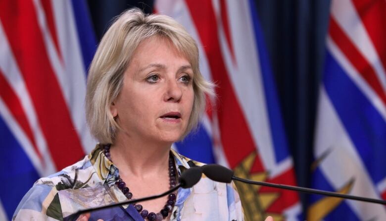 A white woman stands at a podium with the flags of British Columbia in the background.