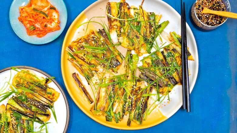 An overhead shot of a plate of pajeon on a bright blue background. A small bowl of sauce, a plate of kimchi and another plate of pajeon surround the main plate. 