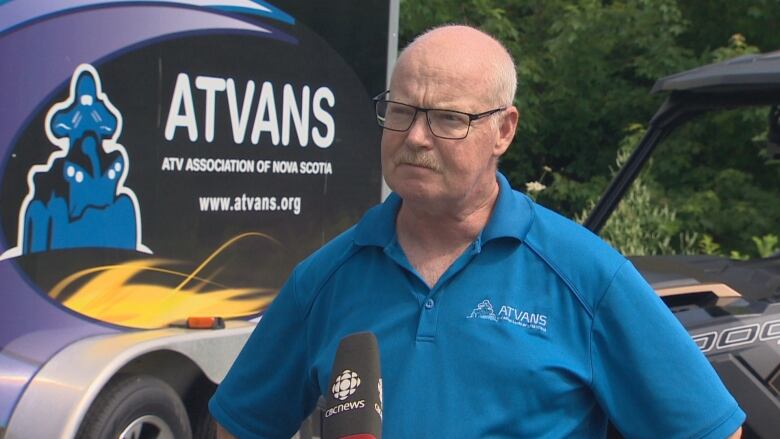 A bald man is wearing rectangular glasses and a blue short-sleeved polo shirt. He is standing in front of a ATV of NS asscociation sign.