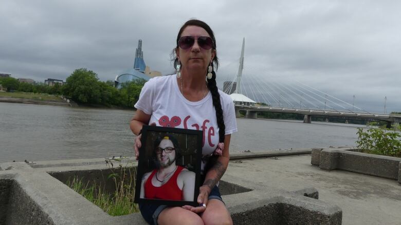 A woman holds a picture of her son while sitting down outside near a river. 