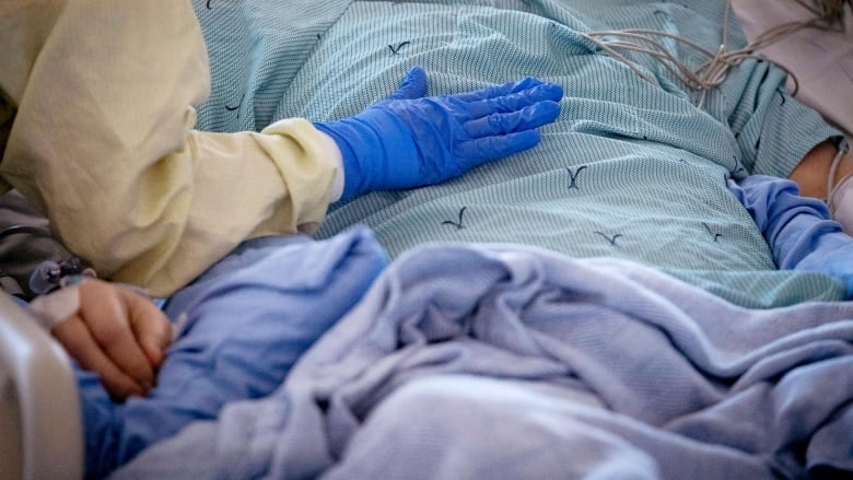 A close up of a person's arm, in a yellow sleeve with a blue latex glove, touching the chest of someone lying on a hospital bed.