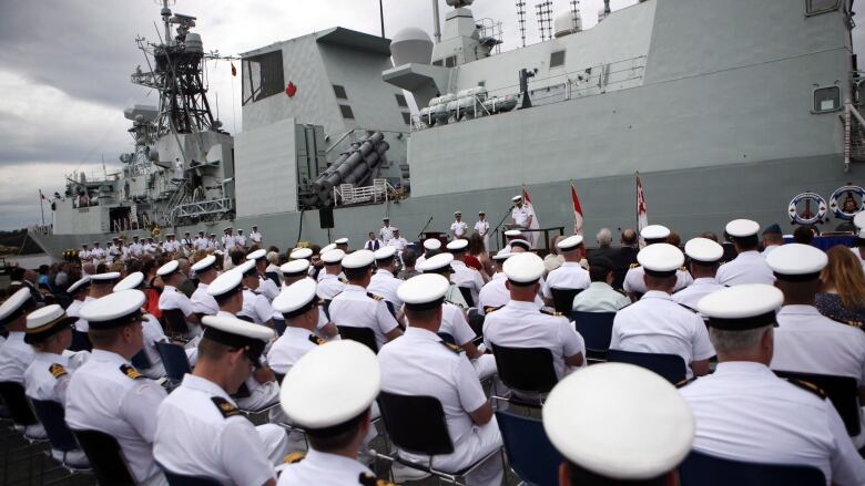 Rows of sailors dressed in white and wearing caps sit before a navy ship.