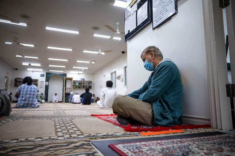 A man sits on a prayer carpet in a Vancouver mosque while wearing a mask. In the background there are other people praying on the floor while spaced out. 