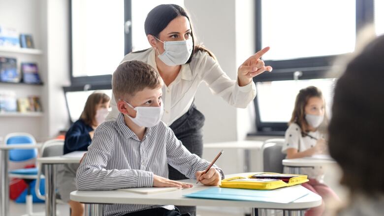 Teacher standing over student pointing to the front of the classroom. Both are wearing facemasks.