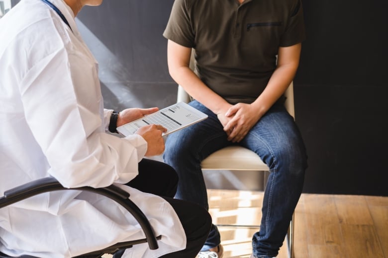 A doctor with a clipboard talks to a patient sitting on a chair.