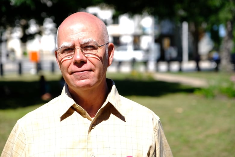 A bald man in a yellow short stands outside in the sunlight.