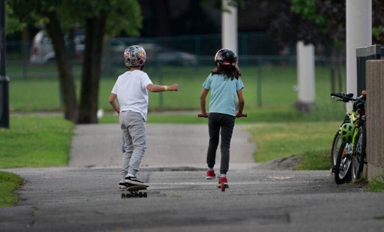 One child on a scooter and another child on a skateboard play in a park.