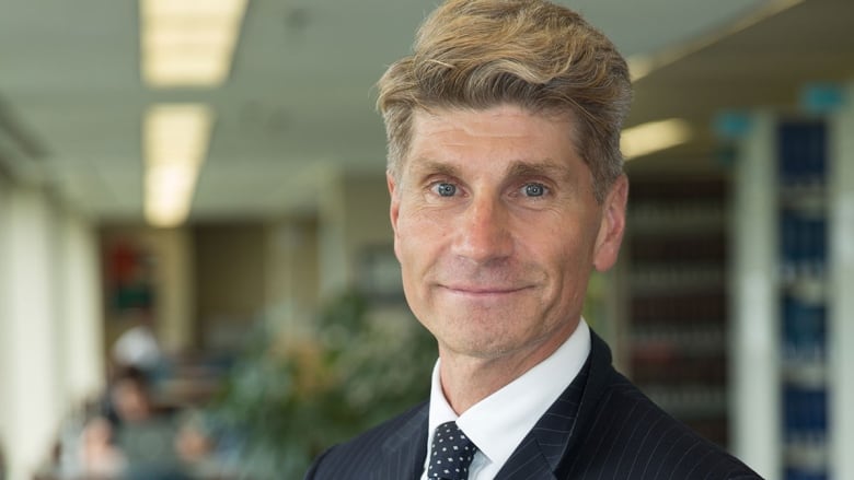 A middle aged man with blonde hair who is wearing a black suit and tie poses for a photo. 