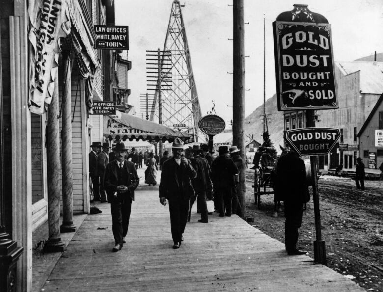 A black-and-white historic photo shows people walking down a wooden sidewalk in a frontier-style town.