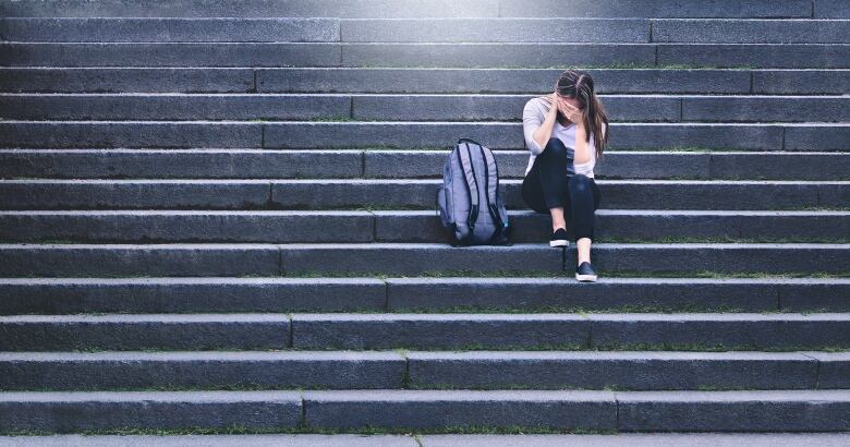 Teenager sitting on stairs with head in hands, backpack on ground next to her.