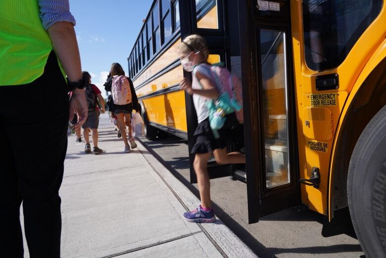 A child in summer clothes steps off a school but and onto a sidewlk while an adult in a safety vest stands by.