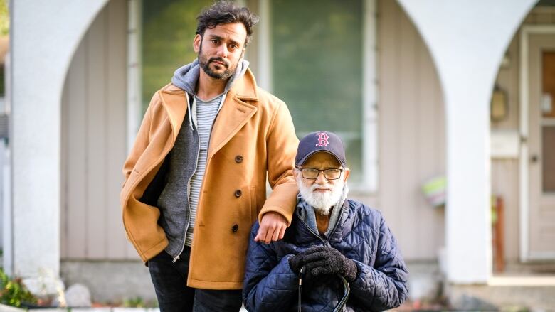 A younger man stands next to an older man seated outside a house.