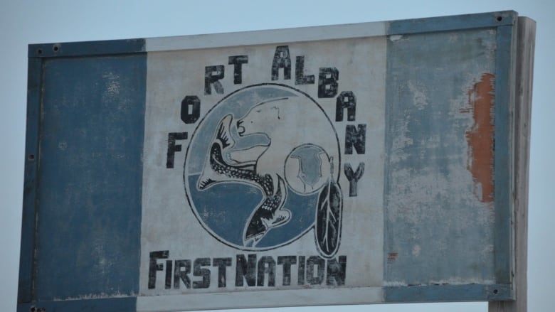 A faded blue and white wooden sign reads 'Fort Albany First Nation' with a picture of a polar bear and a fish