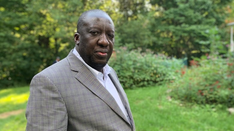 A portrait of a man wearing a blazer and collared shirt, holding a briefcase under his arm, with trees and shrubs in the background. 