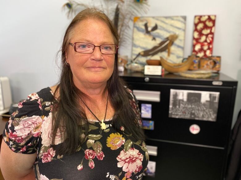 Woman in glasses and floral dress stands in front of a file cabinet on top of which are various pieces of Indigenous art. 