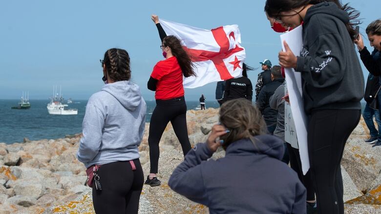 People stand on rocks overlooking the ocean, one of them holding a red and white flag.