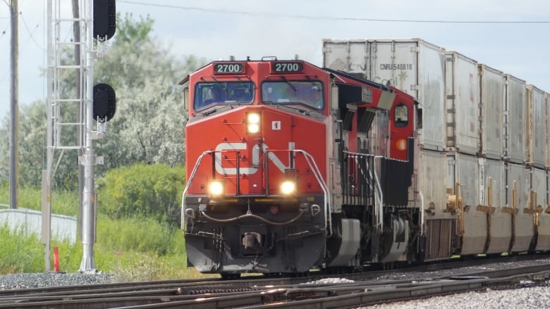 A red train, with 'CN' painted in white on front, is carrying freight cars on a railway.