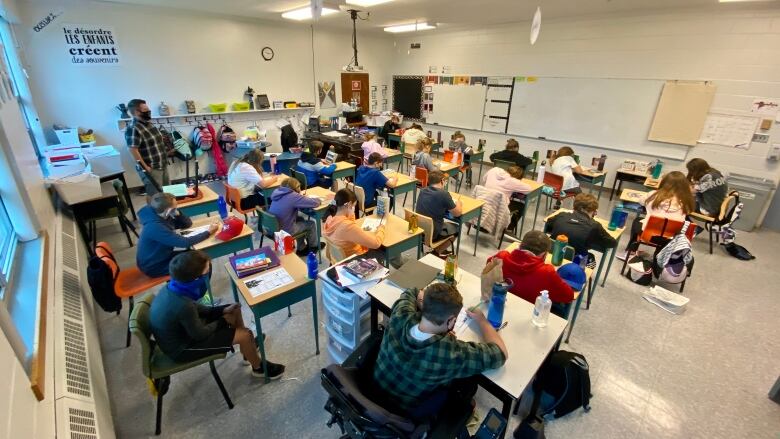 Students and a teacher, all wearing masks, sit in a classroom.