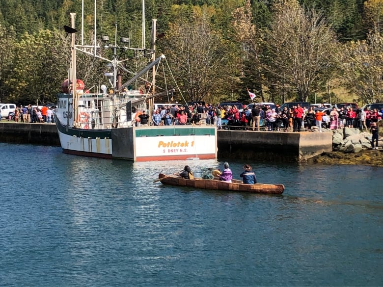 People in a canoe paddle past a large fishing boat tied up in a canal lined with people.