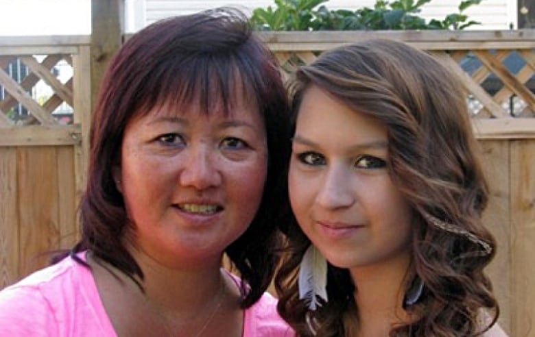 A middle-aged woman with black hair stands next to a teenage girl with long brown hair and feather earrings. Their heads are close together and they are smiling while standing in a yard with a lattice fence in the background.