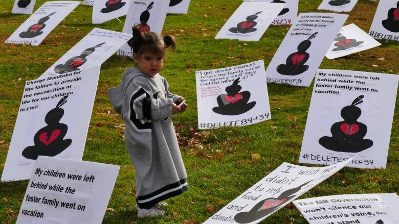 A child in a long grey sweater stands amid a number of signs laid out on the grass. 