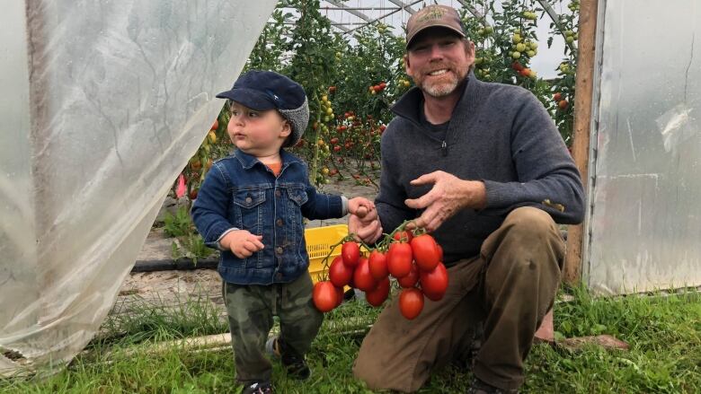 A man holds his son in their greenhouse, the man holds tomatoes in his hand